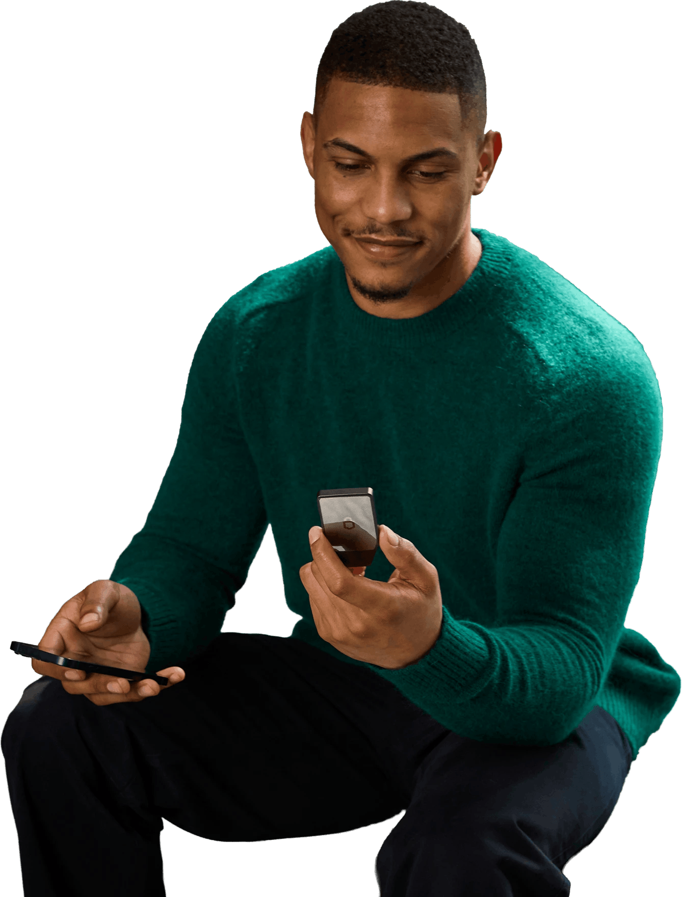 Seated man in a green sweater holding two smartphones against a black background.