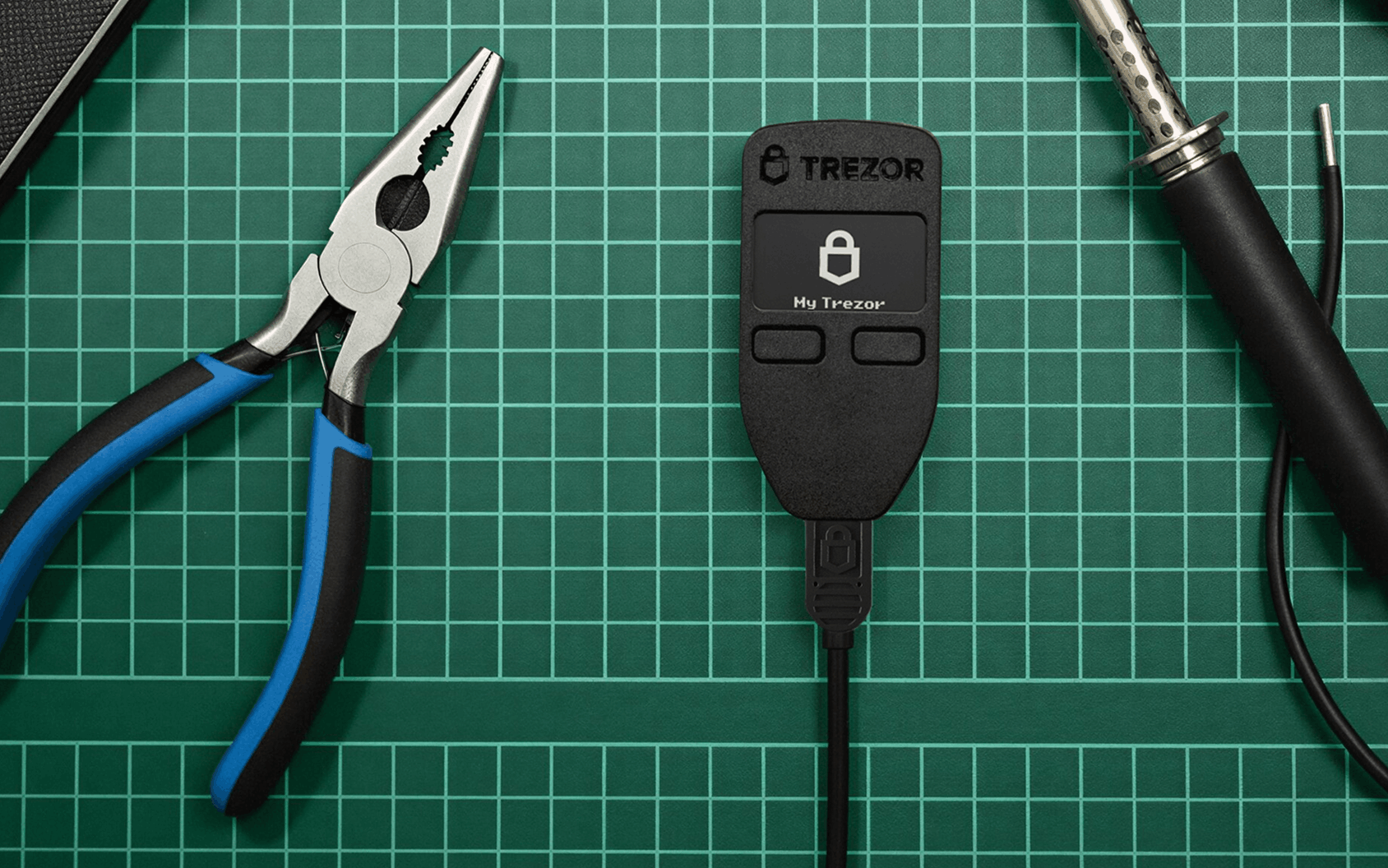 Trezor hardware wallet between blue-handled pliers and a soldering iron on a green gridded cutting mat.