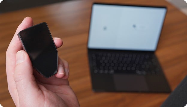 Hand holding a black smartphone in front of an open blurred laptop on a wooden table.