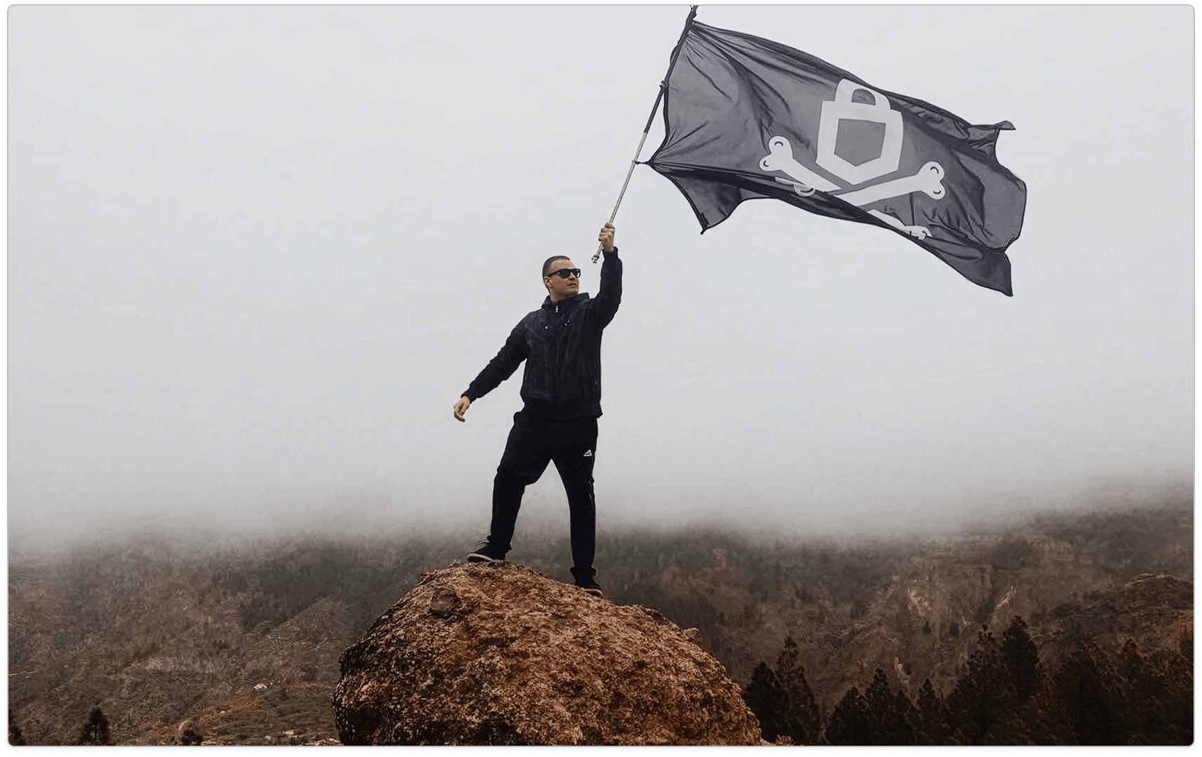 Man standing on a rocky summit holding a large black emblem flag above a hazy forested valley.
